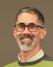 A male adult, with grey glasses, black and white short hair, a white and black goatee, and wearing a green collared shirt beneath a green and grey sweater